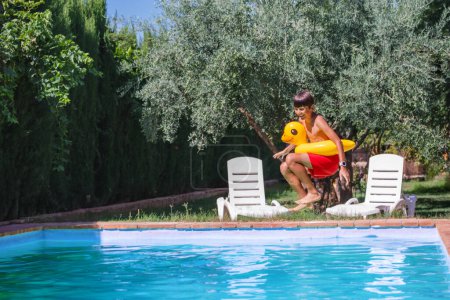 Téléchargez les photos : Heureux adolescent profiter des vacances avec un sourire rayonnant portant maillots de bain cramoisis courses pour plonger dans une piscine, tenir jouet d'eau jaune, jardin verdoyant sur fond - en image libre de droit