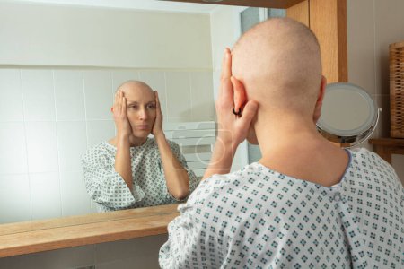 Téléchargez les photos : Une femme chauve en robe d'hôpital examine son reflet dans un miroir de salle de bain - en image libre de droit
