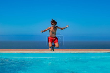 Foto de Feliz adolescente bronceado con pantalones cortos de natación rojos está saltando en las aguas turquesas de una piscina, con el mar como telón de fondo, concepto de vacaciones frescas - Imagen libre de derechos