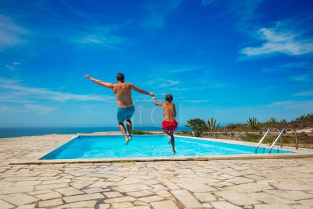 Téléchargez les photos : Une piscine extérieure avec vue sur l'océan est le cadre pour deux personnes, papa et fils plongeant joyeusement dans l'eau bleue chatoyante sous le soleil, vue de derrière - en image libre de droit