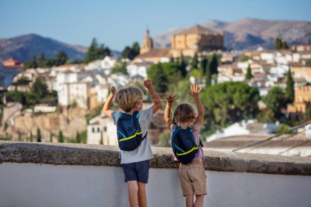 Little travelers stand with backpacks on shoulders look at skyline of morning Ronda during their summer sightseeing trip in Spain