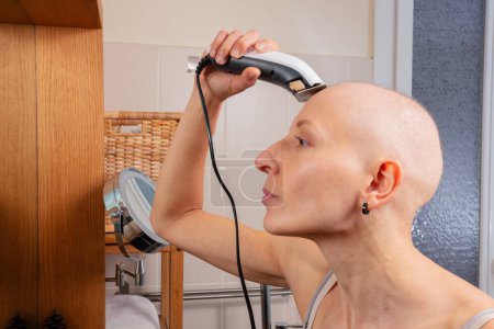 Foto de Mujer sin pelo afeita cuidadosamente su cuero cabelludo con una máquina de afeitar eléctrica, reflejado en el espejo del baño, vista de perfil - Imagen libre de derechos