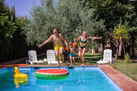 Foto de Niño acompañado por dos adultos experimenta la emoción de sumergirse en una piscina en un día cálido y luminoso, con sillas junto a la piscina y juguetes flotantes cerca. - Imagen libre de derechos