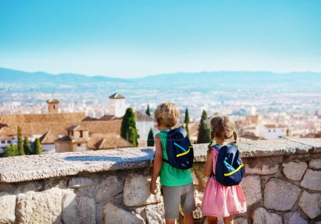 Foto de Niños con mochila, niño y niña de pie en el mirador disfrutan de la vista del antiguo palacio de la Alhambra en su viaje de verano a Granada, España - Imagen libre de derechos
