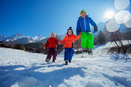 Foto de Niños felices, dos chicos. niña pequeña saltar y correr en la ladera de la montaña soleada en el resort alpino - Imagen libre de derechos