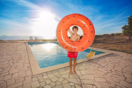 Photo for A happy boy in red swim trunks stands by a pool holding an orange inflatable ring on sunset light at summer resort - Royalty Free Image