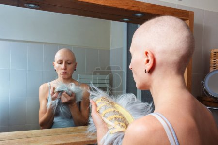 Foto de Mujer calva triste está de pie frente a un espejo, sosteniendo una peluca gris para ponerse, comprometida en la reflexión - Imagen libre de derechos