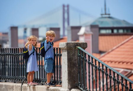 Foto de Dos niños viajeros parados en un mirador en Lisboa, gritan emocionados con mochilas en hombros, puentes y tejados en el fondo - Imagen libre de derechos