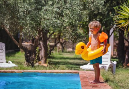 Foto de Un niño sonriendo, sosteniendo un pato amarillo inflable junto a una piscina, con olivos en el fondo, listo para bucear - Imagen libre de derechos
