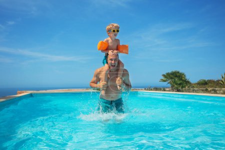 Téléchargez les photos : Un garçon souriant perché sur les épaules d'un nageur masculin, tous deux profitant des eaux fraîches d'une piscine extérieure avec une toile de fond ensoleillée - en image libre de droit