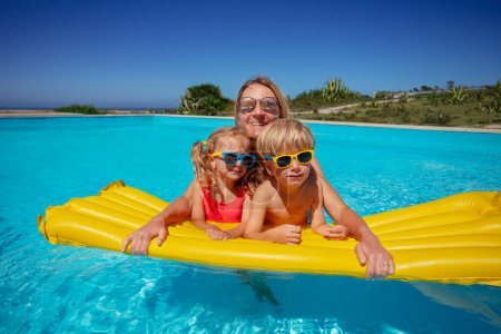 Téléchargez les photos : Trois personnes, un adulte et deux enfants, profitant d'une journée ensoleillée dans une piscine relaxante sur un grand flotteur gonflable jaune - en image libre de droit