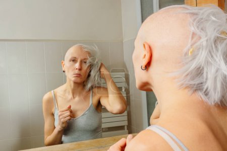 Foto de Contemplando su apariencia, la triste joven con la cabeza afeitada sostiene la peluca de color ceniza mientras se enfrenta a un espejo. - Imagen libre de derechos