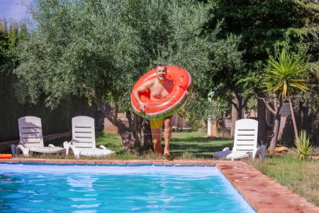 Foto de Un hombre alegre con un anillo inflable colorido se prepara para zambullirse en las aguas cristalinas de una piscina, fijada contra un telón de fondo de árboles y del follaje en su viaje de vacaciones - Imagen libre de derechos