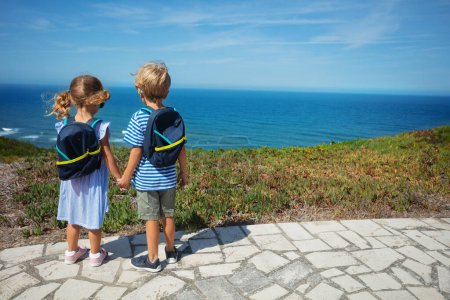 Foto de Niño rubio y niña con mochilas cogidas de la mano admiran el mar desde la colina en el caluroso día de verano en Portugal - Imagen libre de derechos