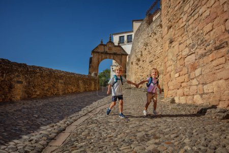 Foto de Niños felices con mochilas corren por las calles pavimentadas de Ronda, disfrutan de sus vacaciones de verano en el sur de España, Europa - Imagen libre de derechos