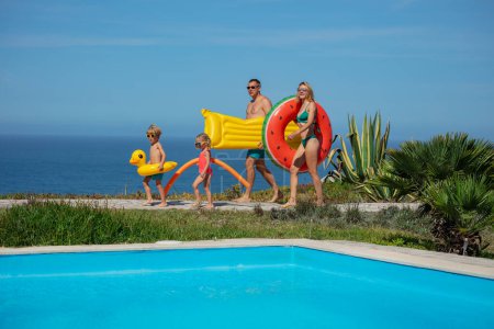 Téléchargez les photos : Près d'une piscine bleu clair, enfants et deux adultes tiennent leurs jouets flottants, prêts à profiter du temps ensoleillé, avec la mer derrière eux - en image libre de droit