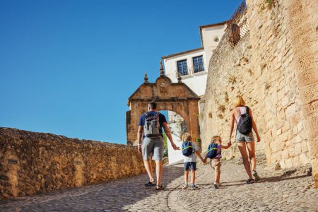 Téléchargez les photos : Famille de touristes avec deux enfants marcher dans la vieille partie de Ronda, aller vers Arco de Felipe V, profiter de leurs vacances d'été dans le sud de l'Espagne, vue de derrière - en image libre de droit