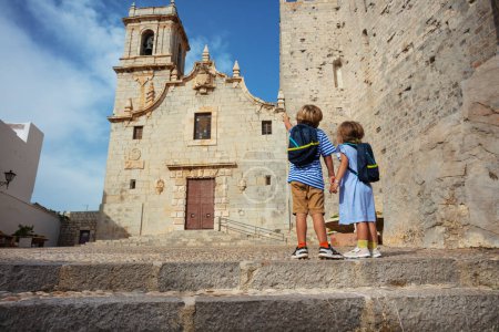 Téléchargez les photos : Les petits voyageurs font du tourisme en Espagne La vieille ville de Peniscola se tient devant l'église de l'Ermitana pointe des doigts au clocher par une chaude journée ensoleillée, vue de derrière - en image libre de droit