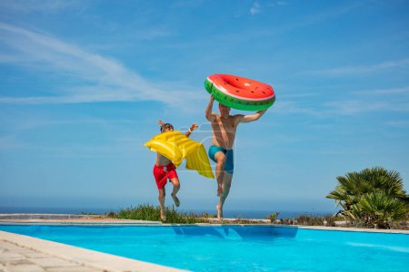 Foto de Dos personas, padre y su hijo con flotadores inflables junto a una piscina bajo un cielo azul claro, listos para bucear - Imagen libre de derechos