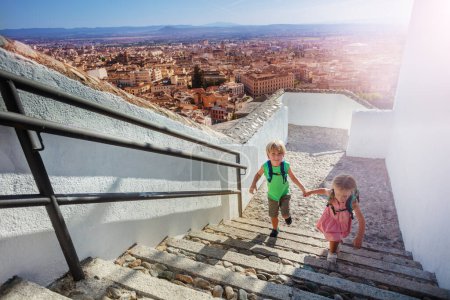 Foto de Dos niños con mochilas suben colina arriba alcanzando mirador para admirar el antiguo palacio de la Alhambra en su viaje de verano a Granada, España - Imagen libre de derechos