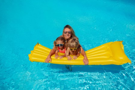Foto de Feliz mamá y dos niños alegres, niño, niña, flotando en aguas azules de la piscina en un colchón amarillo vibrante - Imagen libre de derechos