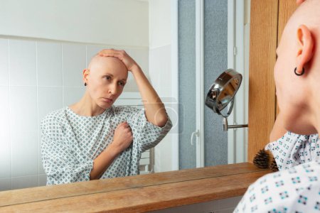 Téléchargez les photos : Reflétant profondément, un patient sans cheveux se regarde dans le miroir de la salle de bain, portant une robe d'hôpital - en image libre de droit