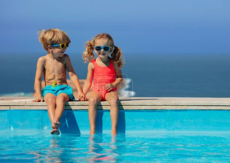 Foto de Dos niños alegres sentados en el borde de una piscina con gafas de sol de colores y trajes de baño con el océano en el fondo - Imagen libre de derechos