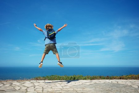 Foto de Niño rubio feliz con mochila y camiseta marina saltar alto emocionado de ver el océano en el día caluroso de verano - Imagen libre de derechos