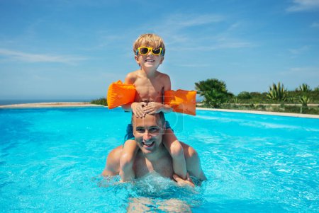 Foto de Un hombre adulto lleva a un niño sobre sus hombros mientras disfruta de un baño en una piscina, rodeado por un cielo despejado - Imagen libre de derechos