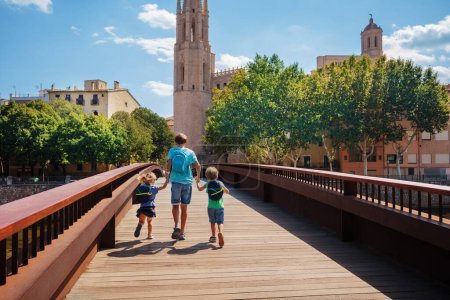 Foto de Tres niños viajeros con mochilas cruzando el puente de Sant Feliu en el centro de Girona en su visita turística de verano en España vista desde atrás - Imagen libre de derechos