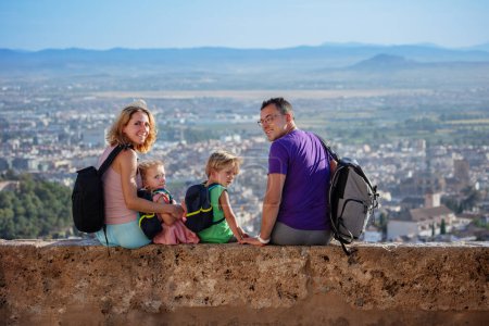 Foto de Familia con niños se sientan en la pared del mirador y vuelven a la cámara emocionados de ver el antiguo palacio de la Alhambra en su viaje de verano a Granada, España - Imagen libre de derechos