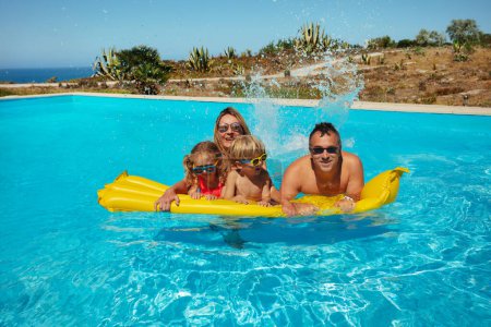 Foto de Dos adultos y niños, todos con gafas de sol, se divierten salpicando en una piscina de color amarillo brillante flotar bajo el sol, con un cielo despejado por encima de ellos sonriendo jugando - Imagen libre de derechos