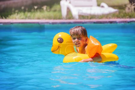 Téléchargez les photos : Un petit garçon jouit d'un temps de jeu aquatique sur un canard gonflable, les bras soutenus par des flotteurs, dans une piscine bleue rafraîchissante dans un jardin - en image libre de droit