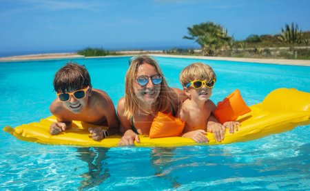 Foto de La madre y sus hijos con gafas de sol comparten un colchón inflable amarillo brillante en aguas cristalinas de la piscina en tiempo soleado caliente - Imagen libre de derechos