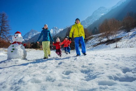 Foto de Familia joven con niños pequeños correr y saltar en el campo nevado sobre picos de montaña soleados, disfrutar de vacaciones de invierno - Imagen libre de derechos