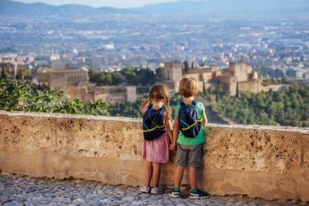 Foto de Pequeños mochileros parados en el mirador hipnotizados por el antiguo castillo de la Alhambra en su viaje familiar de verano a Granada, España - Imagen libre de derechos