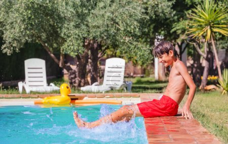 Foto de Joven niño sonriente en bañador rojo chapotea alegremente en una piscina en un día soleado, con follaje verde y sillones en el fondo - Imagen libre de derechos