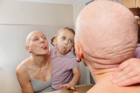 Téléchargez les photos : Une femme chauve combattant le cancer et une jeune fille faisant des grimaces à leur reflet dans le miroir de la salle de bain - en image libre de droit