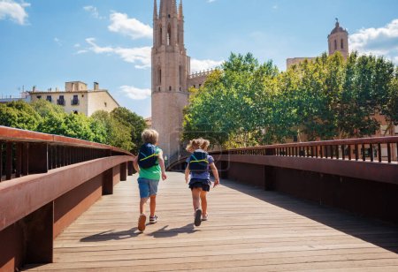 Téléchargez les photos : Garçon et fille avec des sacs à dos courir sur le pont Sant Feliu dans le centre-ville de Gérone lors de leur visite touristique d'été en Espagne, vue de derrière - en image libre de droit