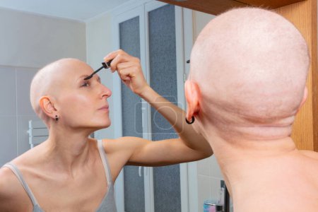 Foto de Chica joven con problemas de salud ponerse maquillaje de ojos mientras mira a su reflejo en un espejo, mantenerse fuerte en la lucha contra el cáncer - Imagen libre de derechos