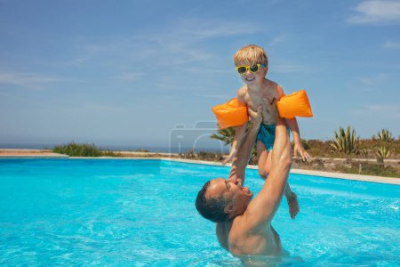 Photo for A young boy with inflatable orange arm bands and sunglasses is joyfully lifted up by an adult male in a sunlit outdoor swimming pool - Royalty Free Image
