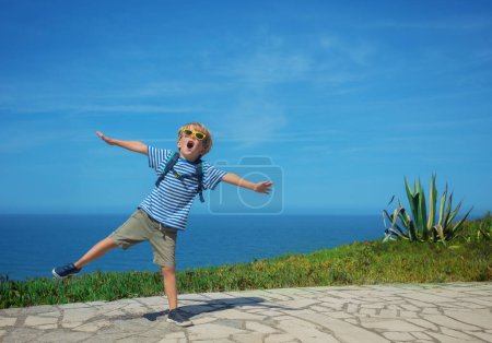 Téléchargez les photos : Garçon blond excité en tenue d'été avec des lunettes de soleil dansant et chantant sur fond de mer - en image libre de droit