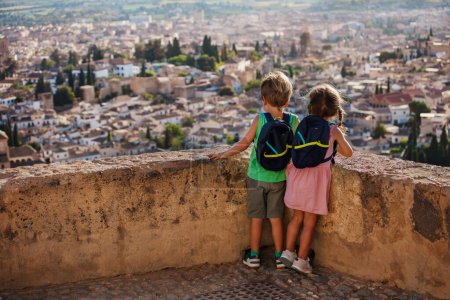 Foto de Hermano y hermana con mochilas de pie en el mirador disfrutan de la antigua ciudadela de la Alhambra en su viaje familiar de verano a Granada, España - Imagen libre de derechos