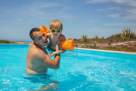 Foto de Un hombre levanta a un niño sonriente, que está equipado con flotantes alas de agua naranja y tonos deportivos, por encima de las aguas cristalinas de una piscina al aire libre - Imagen libre de derechos