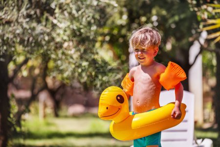 Foto de Niño alegre listo para nadar en su anillo de natación en forma de pato en el borde de una piscina azul claro, con follaje natural detrás - Imagen libre de derechos
