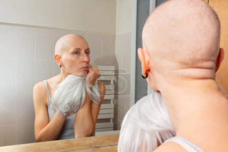 Photo for Sad woman struggling of cancer with a shaved head examines reflection in a bathroom mirror, holding white wig - Royalty Free Image