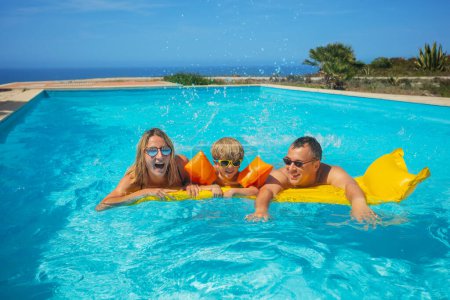 Foto de Un grupo de tres, mamá, papá y un niño con traje de baño experimenta el placer de un refrescante chapuzón en una piscina con un paisaje marino panorámico en el fondo - Imagen libre de derechos