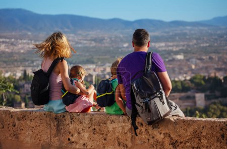 Foto de Turistas con niños se sientan en la pared en el mirador y el descanso, con vistas a la Alhambra antigua ciudadela en su visita a Granada, España - Imagen libre de derechos