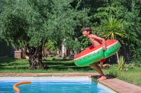 Foto de El muchacho joven que sostiene un anillo inflable de la sandía se prepara para saltar en una piscina en un día soleado, rodeado de vegetación exuberante, concepto de vacaciones - Imagen libre de derechos