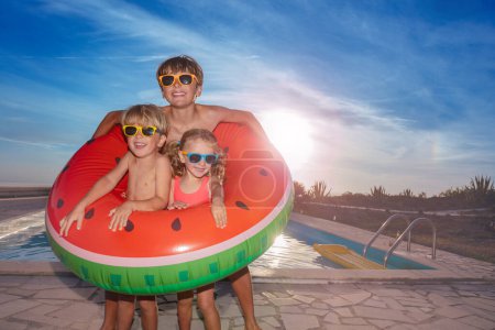 Foto de Dos chicos y una chica en traje de baño. las gafas de sol posan con una sandía grande inflable por una piscina, contra un telón de fondo del sol poniente. - Imagen libre de derechos